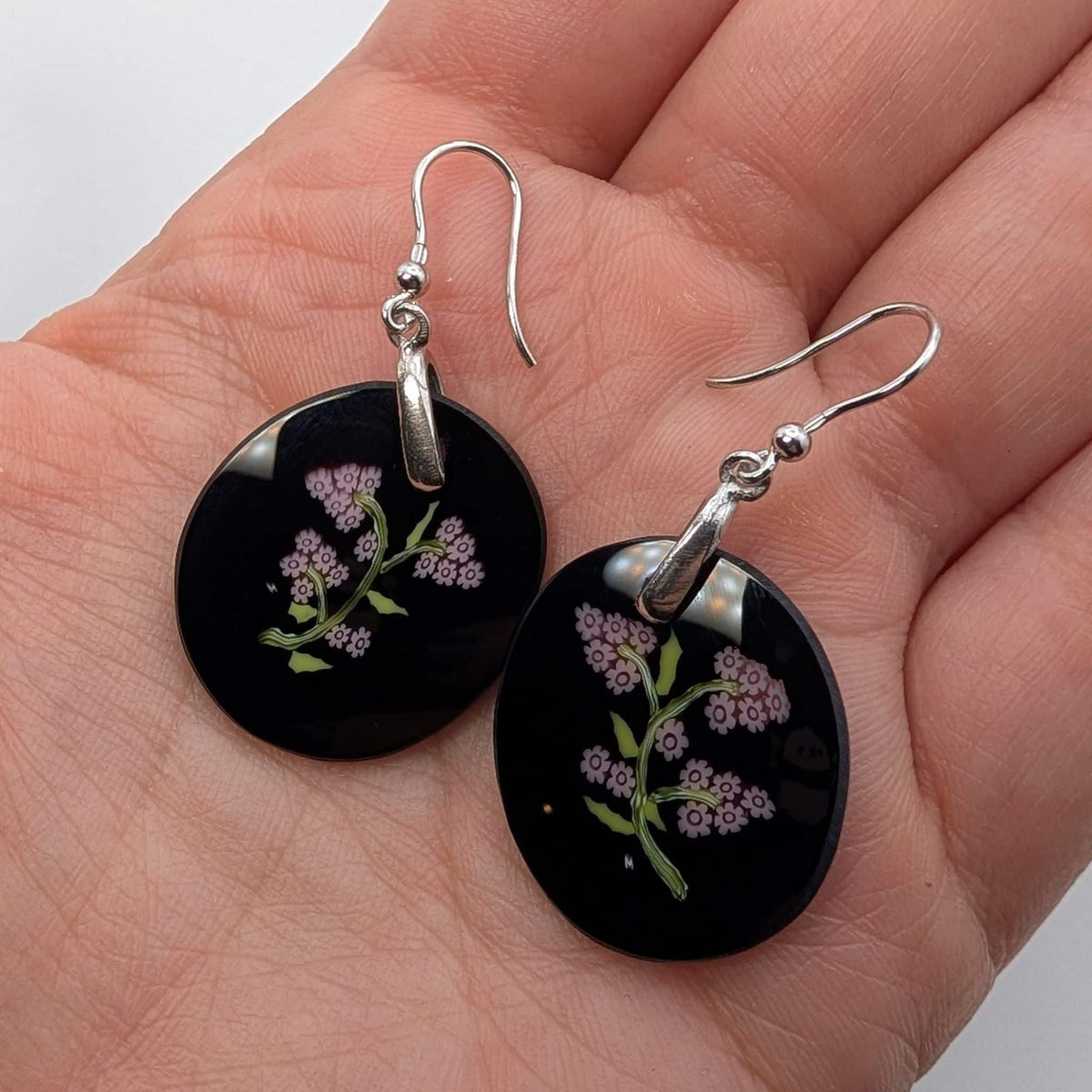 A pair of murano glass round black dangle earrings with silver fishhook wires and pink flowers and green stems, displayed on a person's palm.