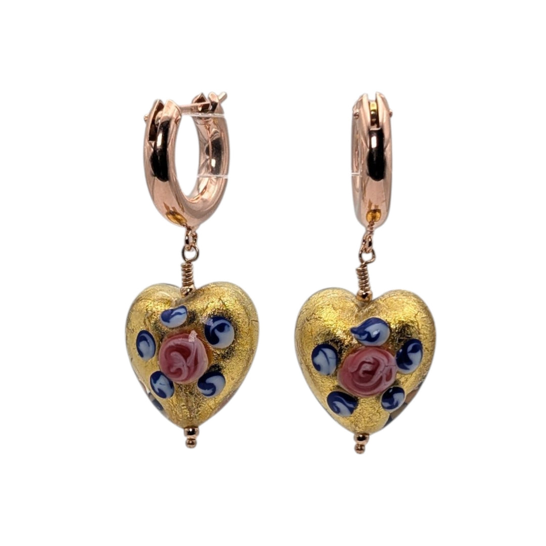 A pair of rose gold huggie hoop earrings with dangling heart charms. The heart charms feature a textured gold background decorated with blue and pink rose motifs, set against a plain white background.