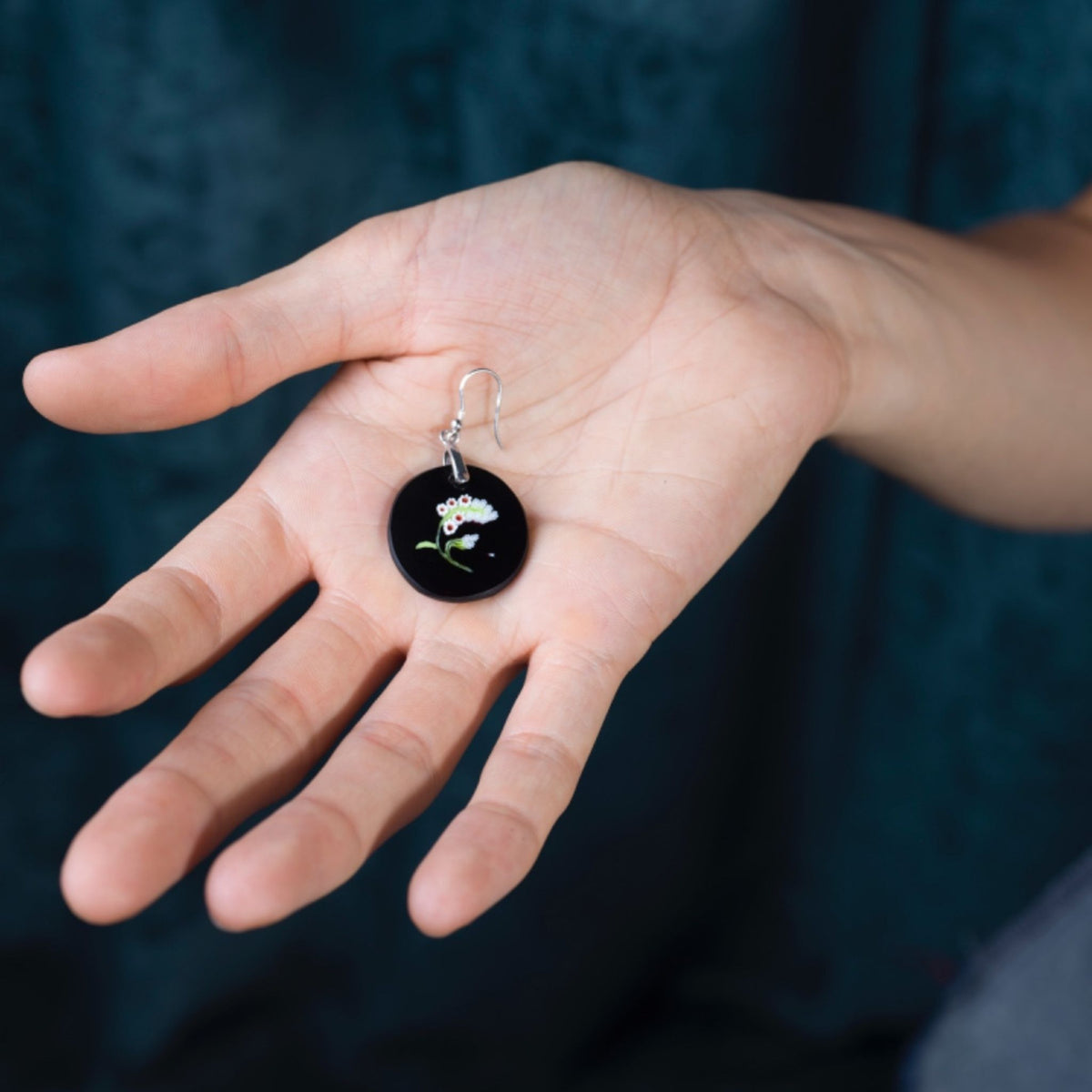 A close-up of a hand holding a small round black murano glass earring with a silver fishhook and a tiny white flower design.