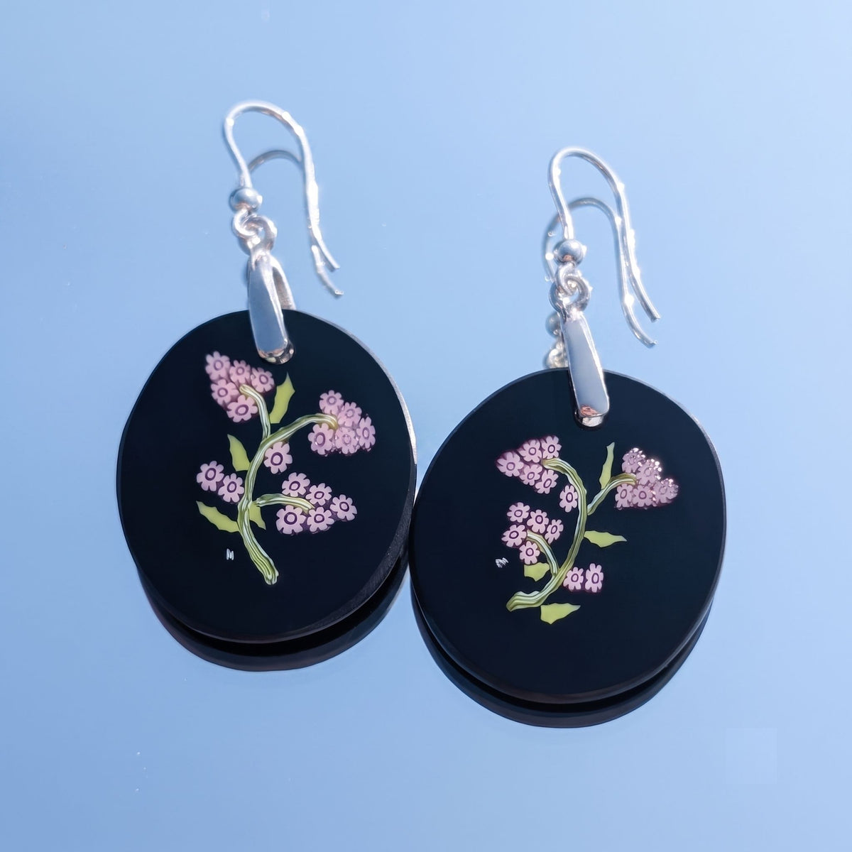 A pair of black circular drop earrings with silver fishhook wires, decorated with murano glass millefiori pink flowers and green stems.