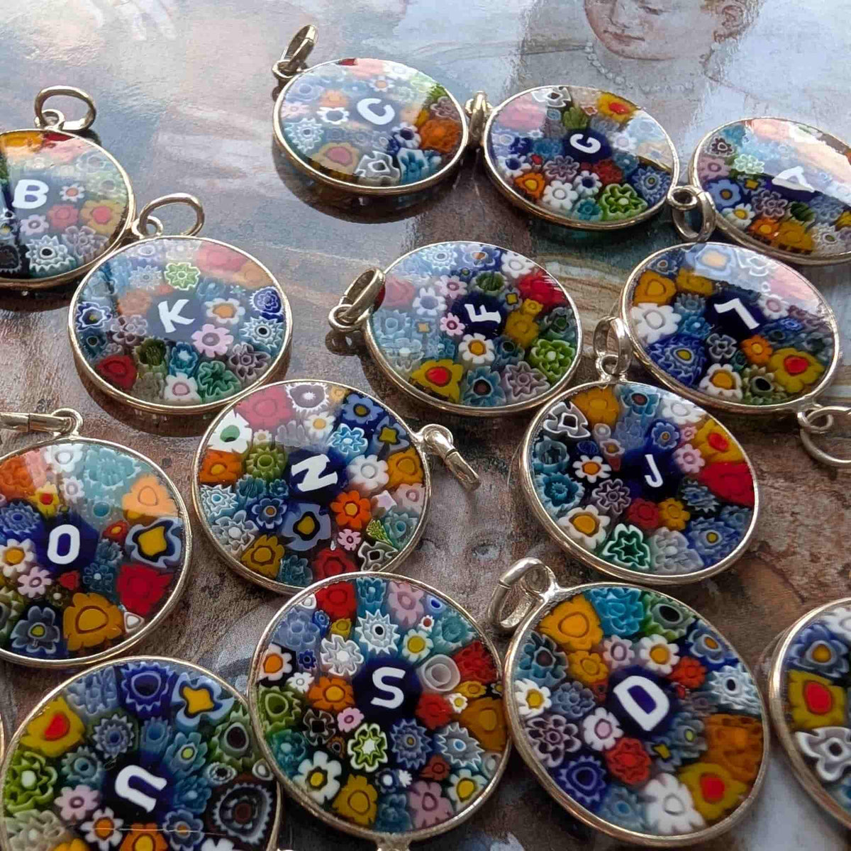 A group of round glass millefiori pendant charms featuring colourful floral patterns and personalised initial letters, displayed on a textured surface.