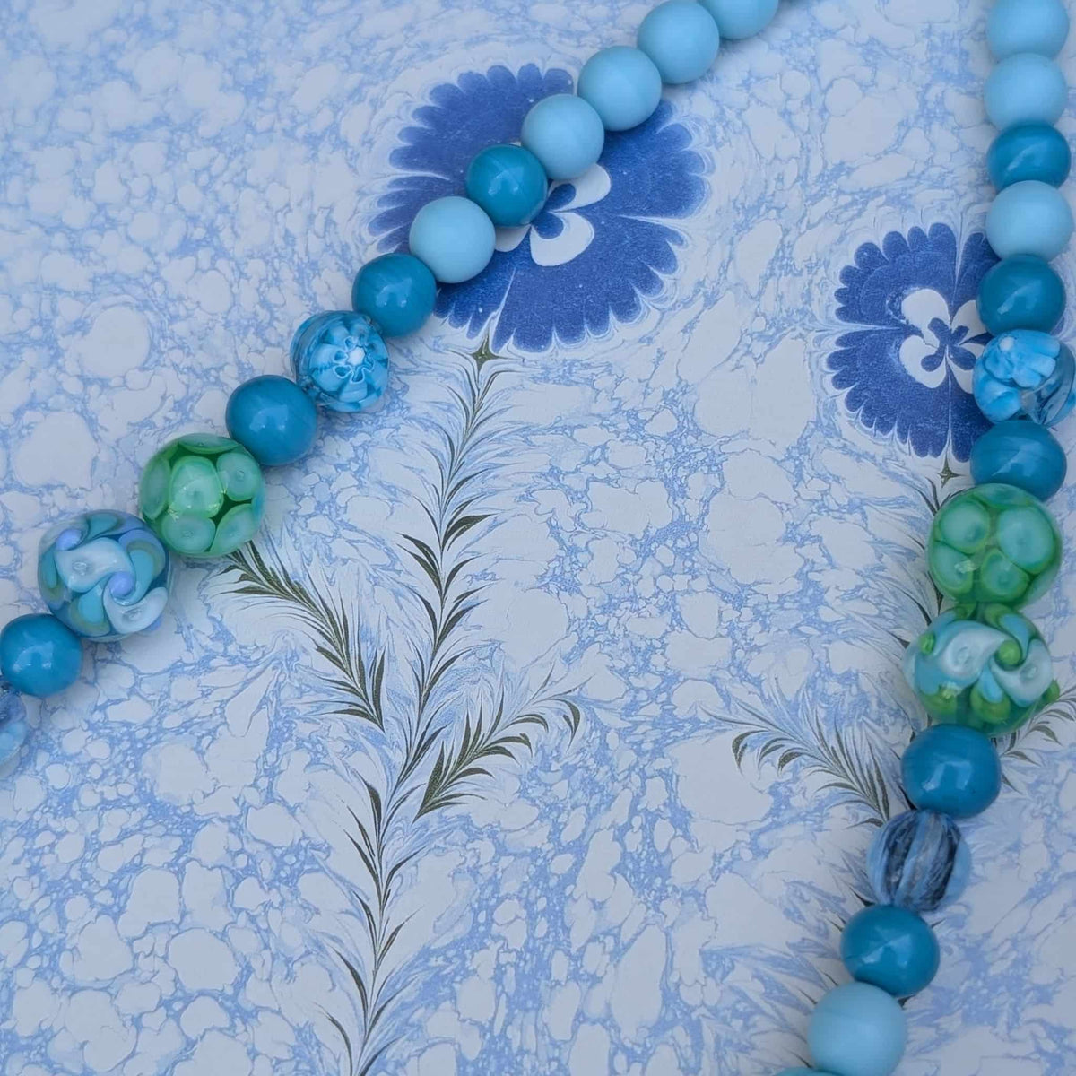 A beaded strand of round glass beads in shades of light blue, medium blue, teal, and green, laid diagonally on a blue and white floral patterned surface.