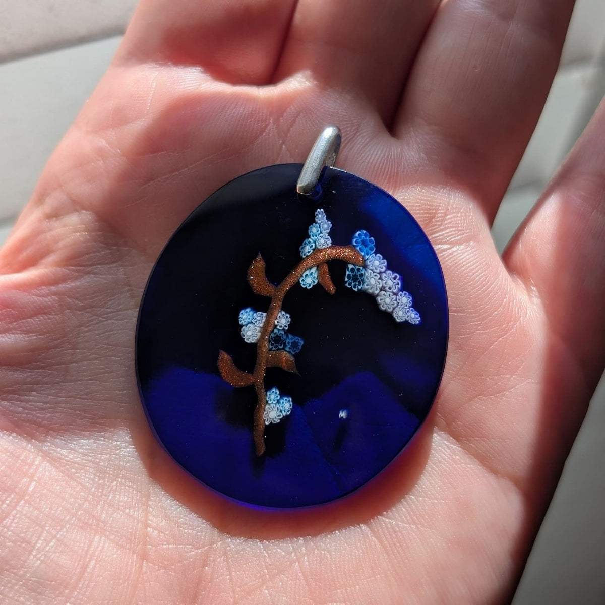A hand holding a round dark blue glass pendant with a silver bail, featuring an artistic design of a brown branch decorated with light blue and white glittery floral clusters.