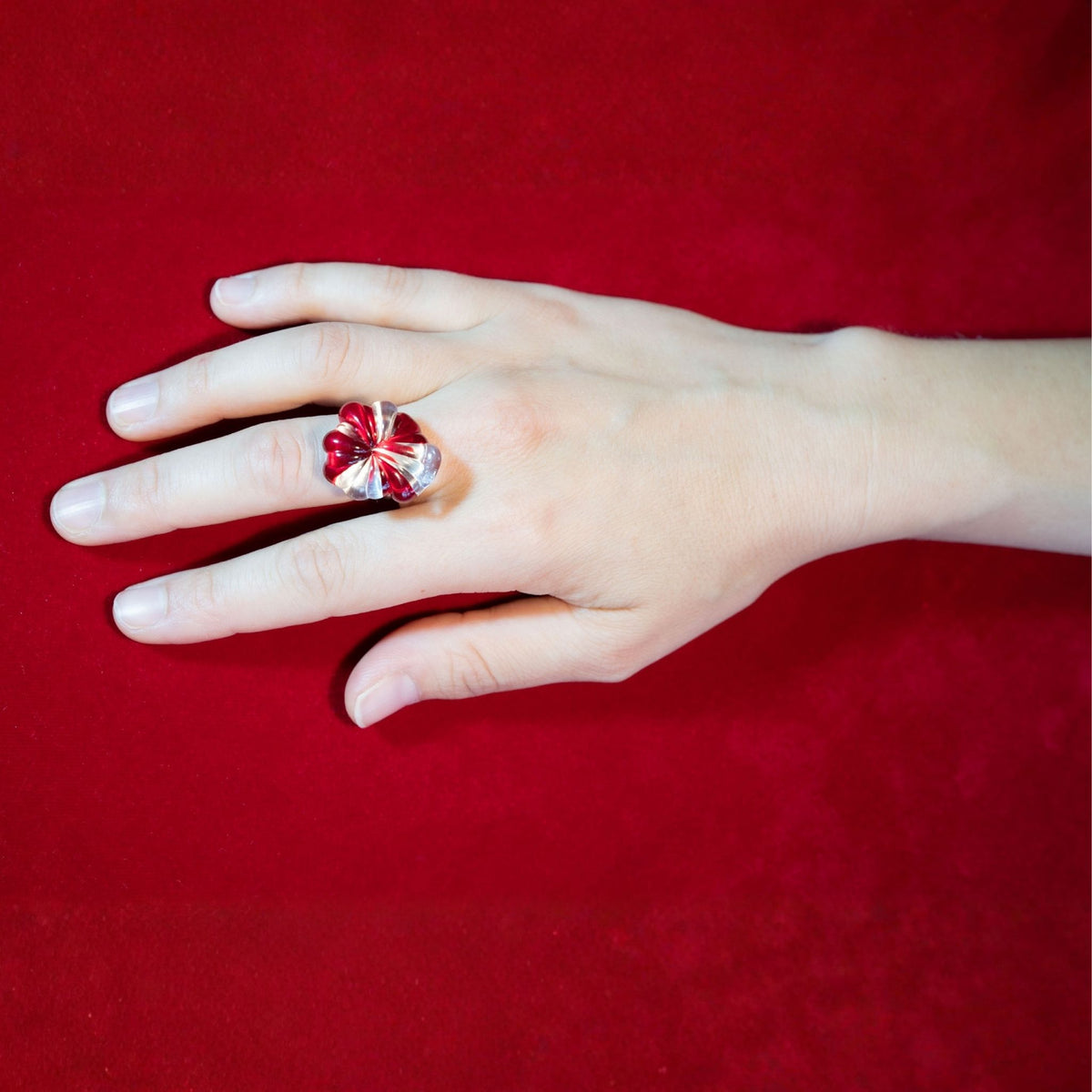 A hand wearing a silver ring with a red and clear glass ring, set against a velvet red background.