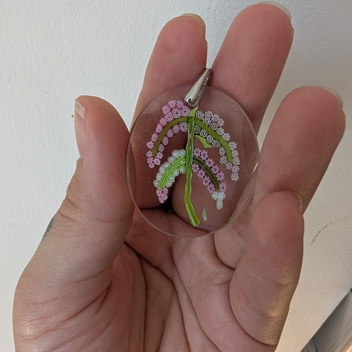 A hand holding a small oval glass clear pendant or charm featuring a delicate floral design in pink, white, and green, with a silver-toned bail at the top.