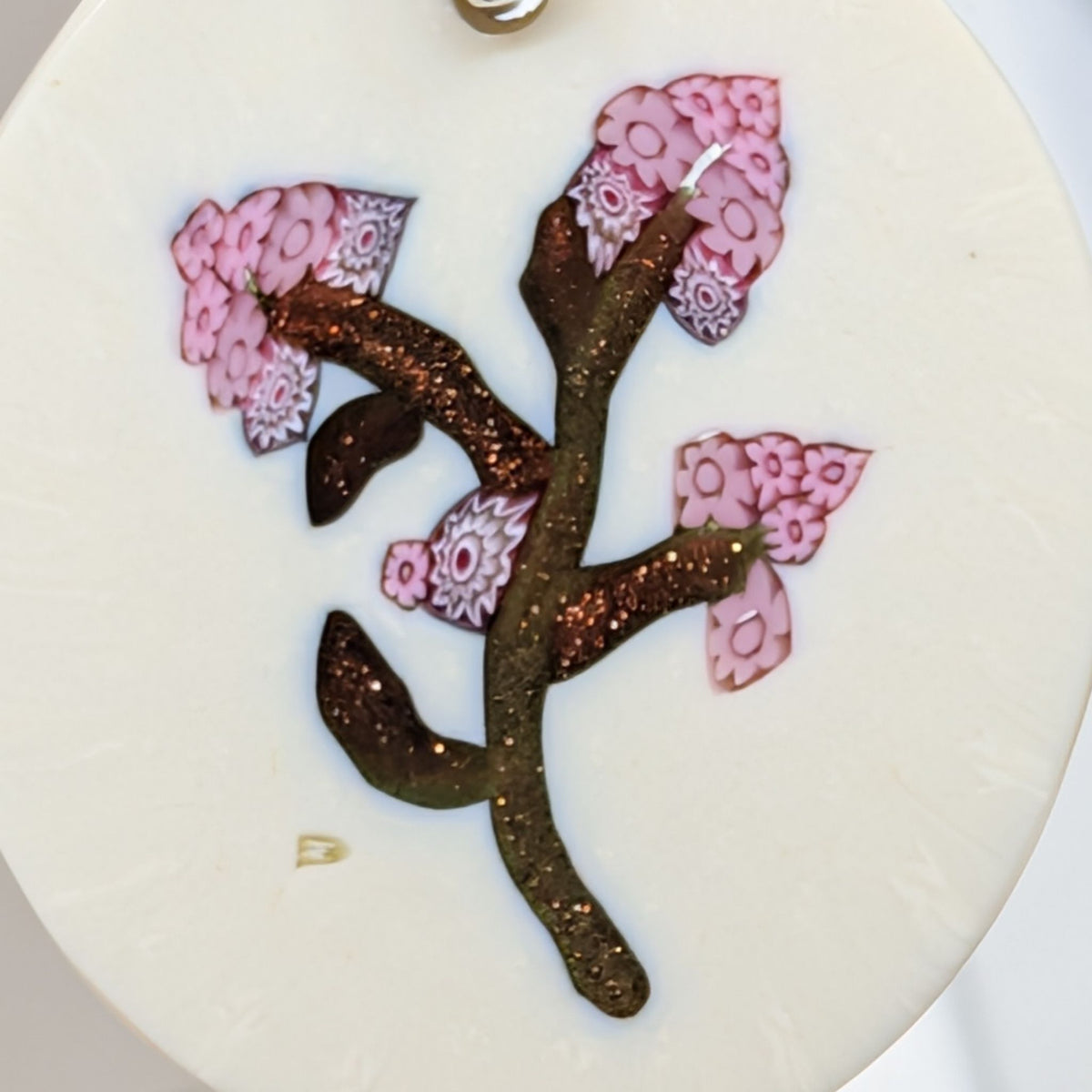 A close-up of a circular white decorative earring millefiori featuring a stylised cherry blossom branch with pink patterned petals and glittery brown leaves.