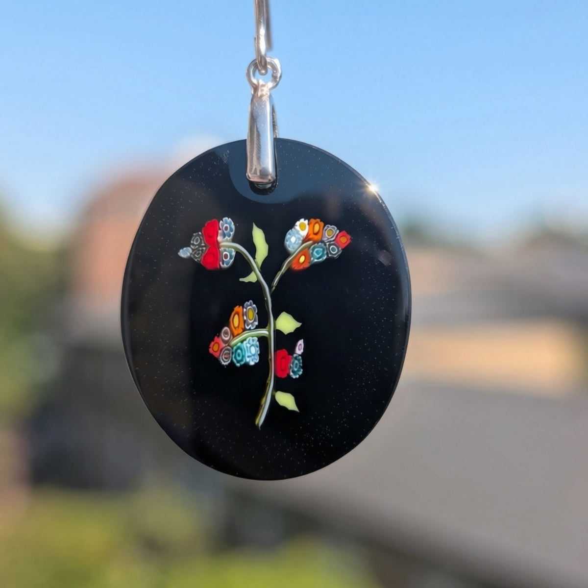 A close-up of a black oval pendant with a colourful glass floral design, hanging from a silver bail against a blurred outdoor background.