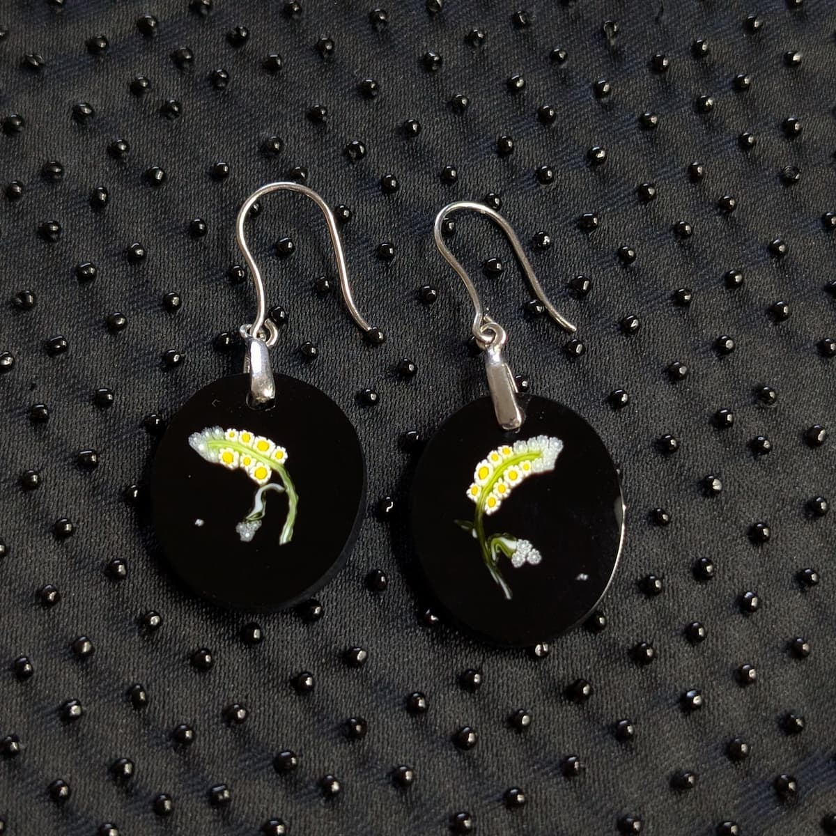 A pair of round black dangle earrings with silver fishhook wires, featuring glass yellow and white flower motif on the front, displayed on a black textured background.