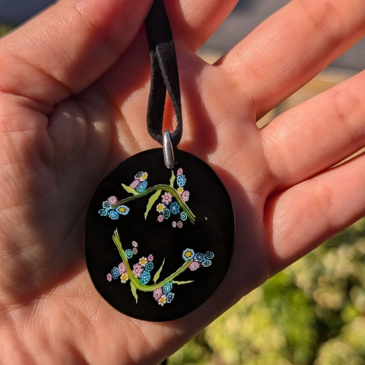A close-up of a hand holding a round black pendant with colorful floral and butterfly designs, attached to a black ribbon, with a blurred outdoor background.