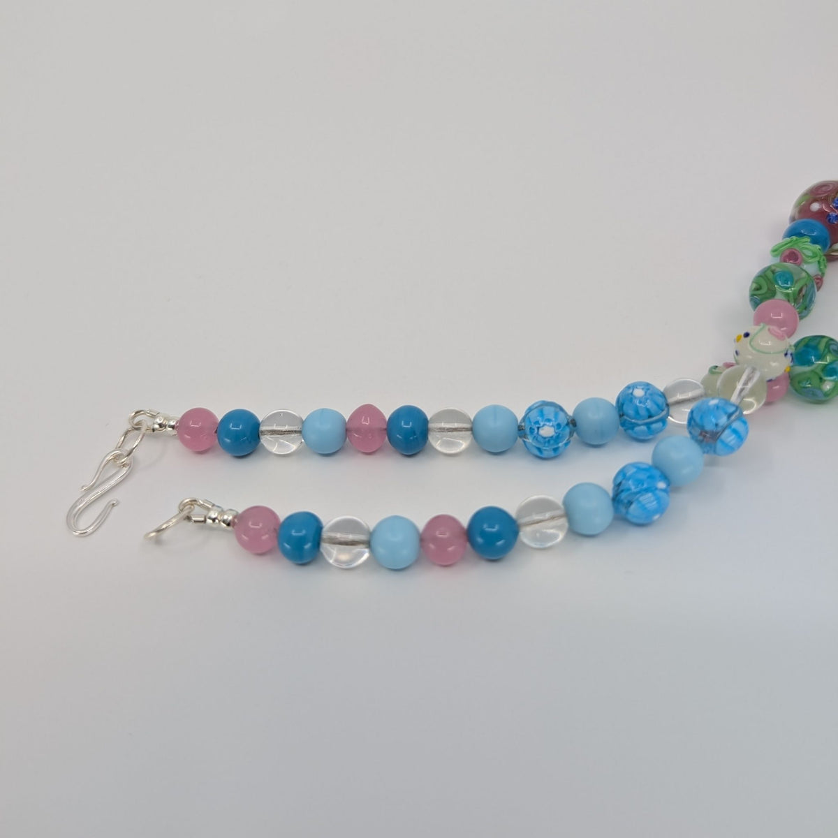 A beaded necklace with alternating light blue, medium blue, pink, and clear faceted beads, displayed on a light gray background.