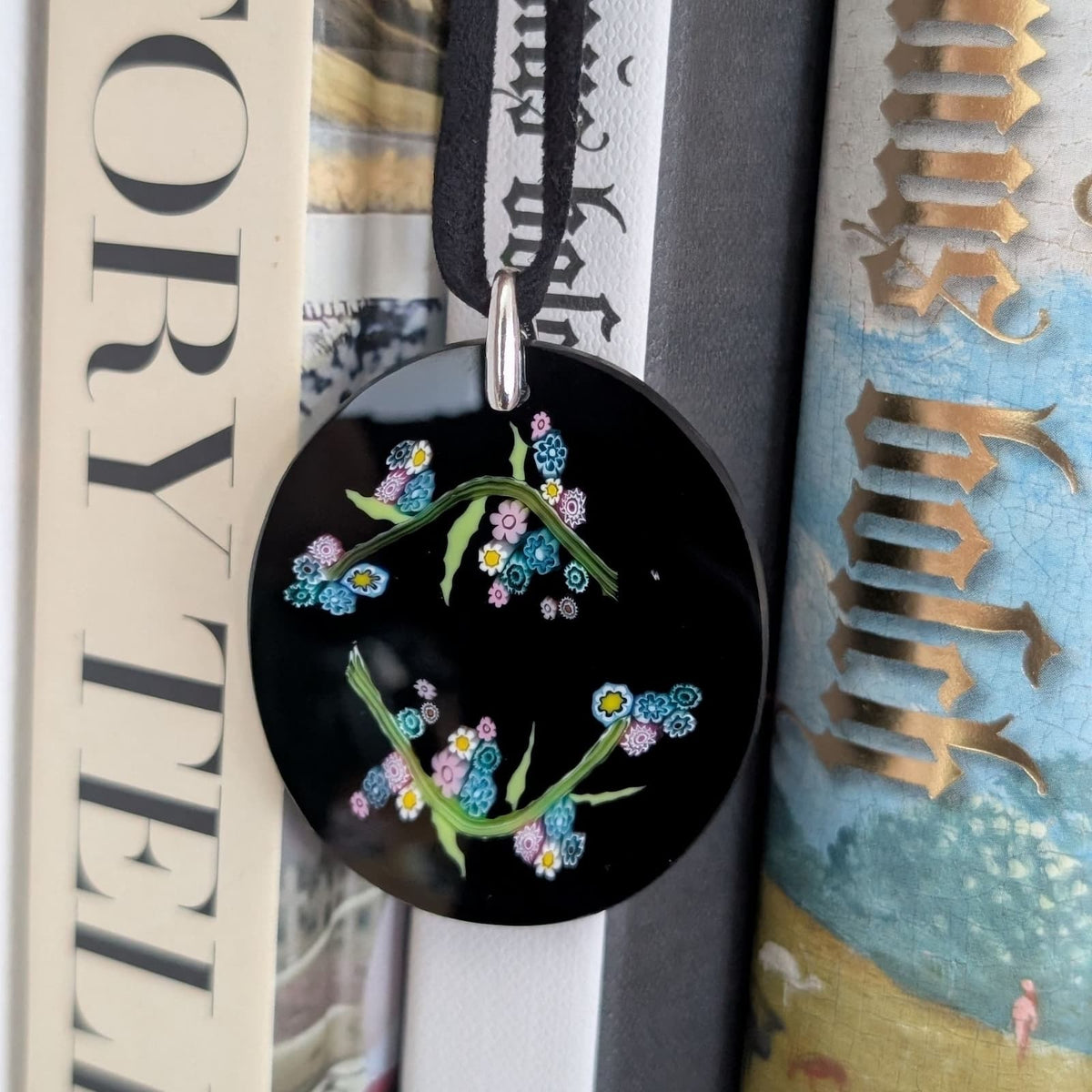 A round black pendant with a silver-tone top loop, decorated with millefiori glass forget-me-not flowers on a black ribbon, hanging between books.