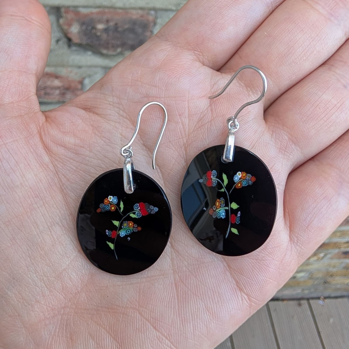 A pair of round black dangle earrings with colorful butterfly and floral motifs, hanging from silver fishhook wires and displayed on an open hand.