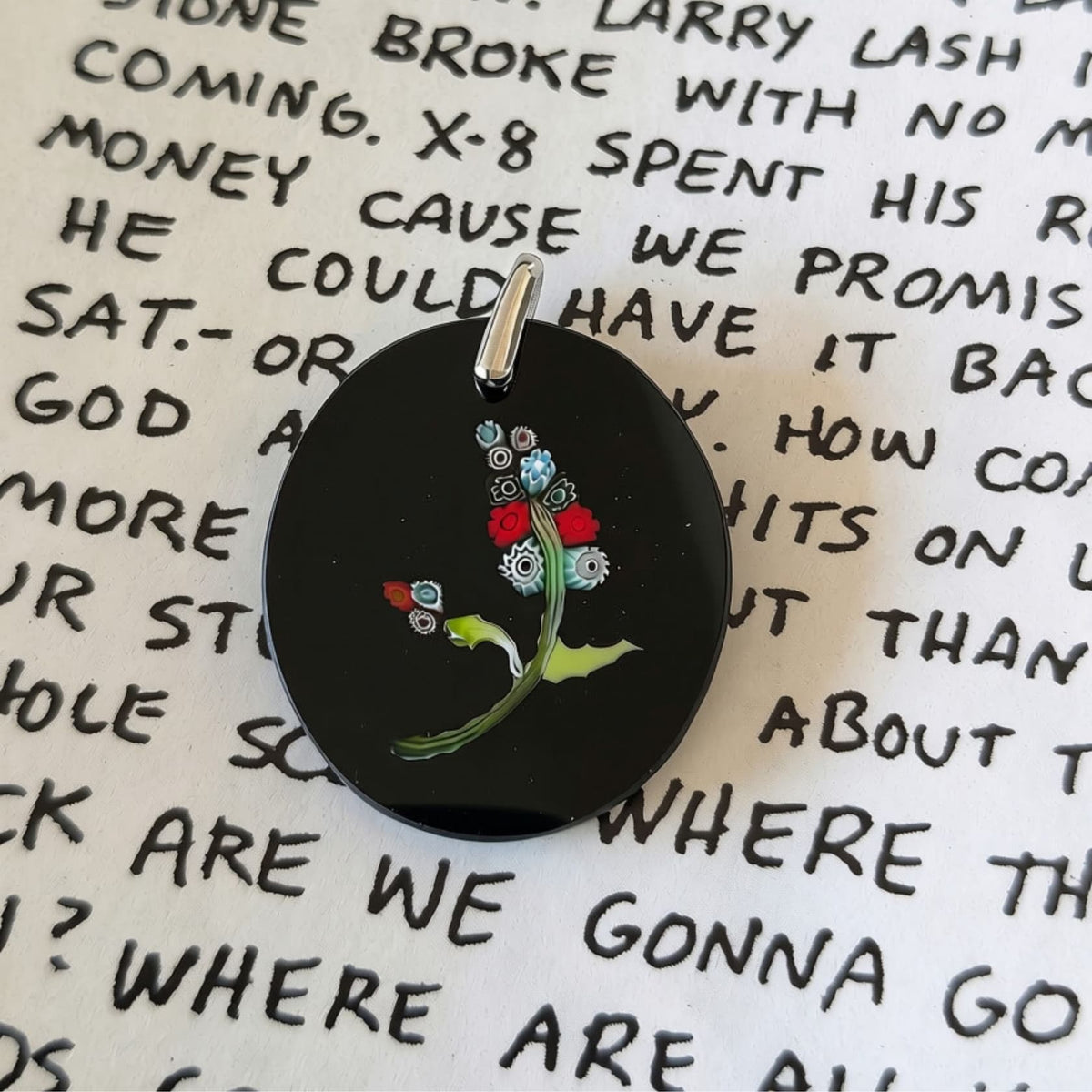 A round black pendant featuring a colorful floral design with a silver bail, resting on a white background printed with handwritten lyrics.