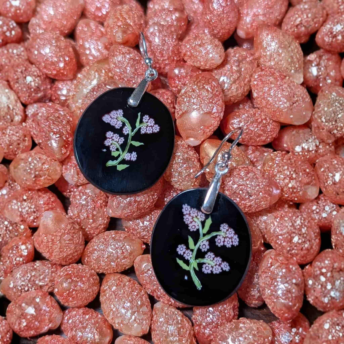 A pair of round black pendant earrings with silver fishhook ear wires, featuring millefiori pink flowers and green stems, displayed on a bed of sparkling peach-coloured beads. all murano glass.