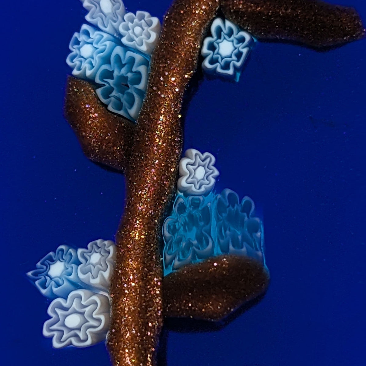 A close-up of a avventurina gold glittery branch or vine with several small white and light blue flower-shaped embellishments attached against a deep blue background.