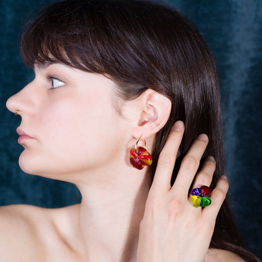 A close-up side profile of a woman wearing a matching pair of colorful flower-shaped drop earrings and a matching flower ring, set against a dark textured blue background.