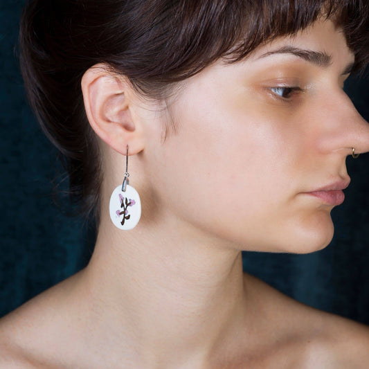 A close-up side profile of a woman wearing a single white oval dangle earring with flowers glass design and a small purple accent, against a dark teal background.