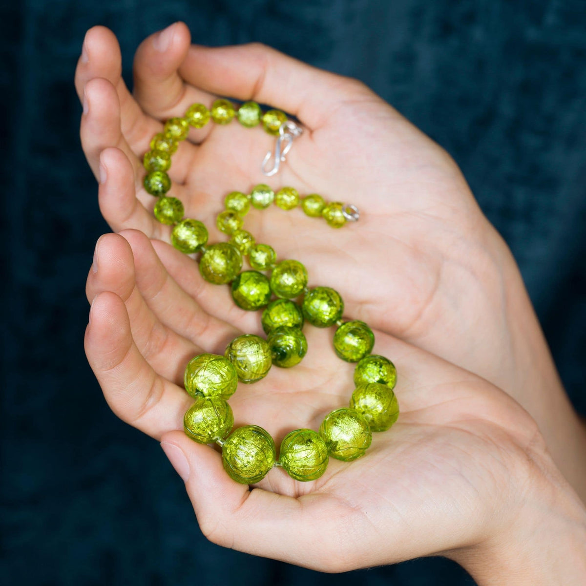 A pair of hands holding a beaded necklace made of shiny, metallic lime green beads with a silver clasp, set against a dark textured background.