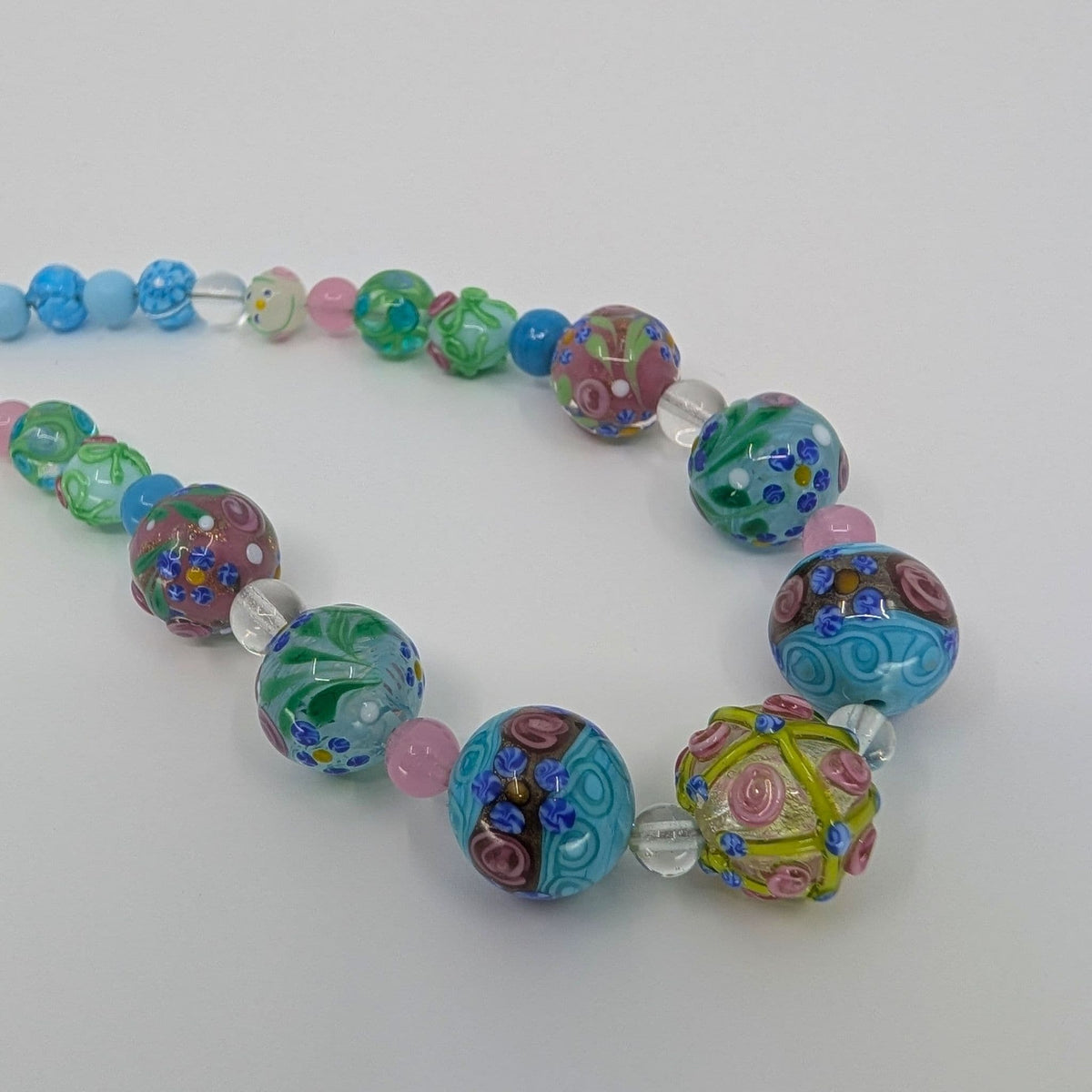 A colorful glass bead necklace featuring various patterned lampwork beads in shades of blue, green, pink, purple, and yellow, accented by clear glass beads, on a plain light gray background.