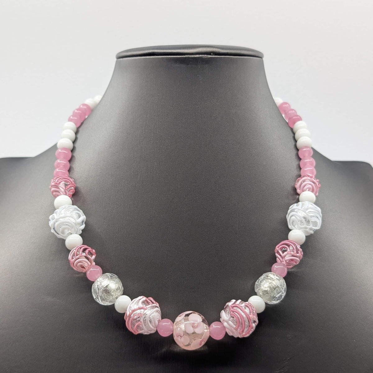 A pink and white beaded necklace displayed on a black mannequin bust. The necklace features a mix of smooth white beads, translucent pink beads, and carved glass beads in pink, white, and clear.