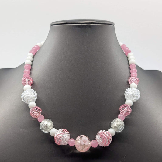 A pink and white beaded necklace displayed on a black mannequin bust. The necklace features a mix of smooth white beads, translucent pink beads, and carved glass beads in pink, white, and clear.