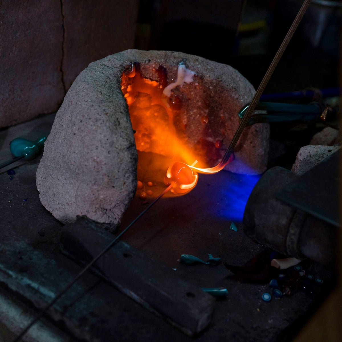 A glassblower's furnace in action, showing a glowing orange-red molten glass bead inside a stone dome, with a metal tool holding a piece of glass near the flame.
