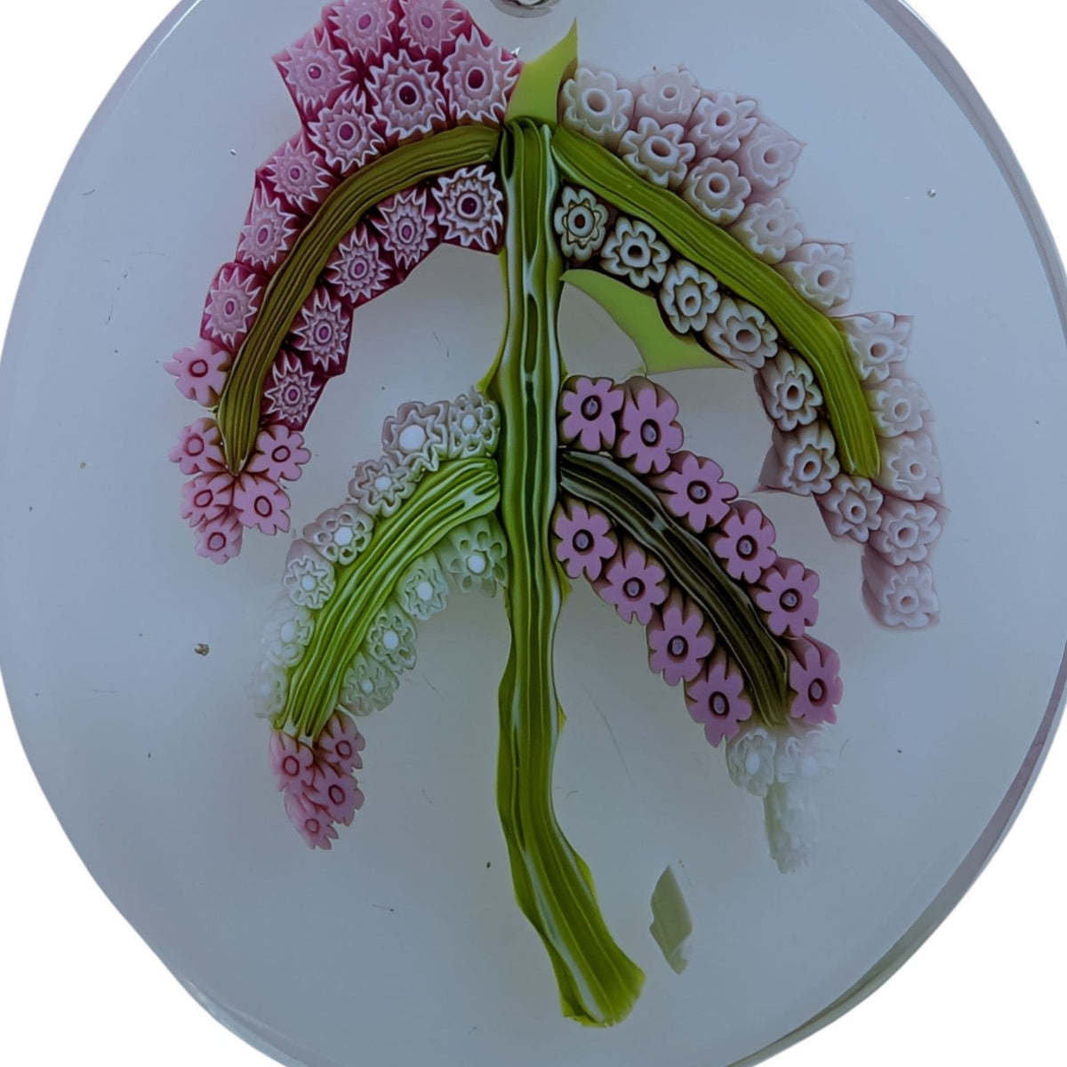 A close-up of a circular glass pendant or ornament featuring a stylized leaf design made from pink, green, and white millefiori or pearly glass petal patterns on a clear glass base.