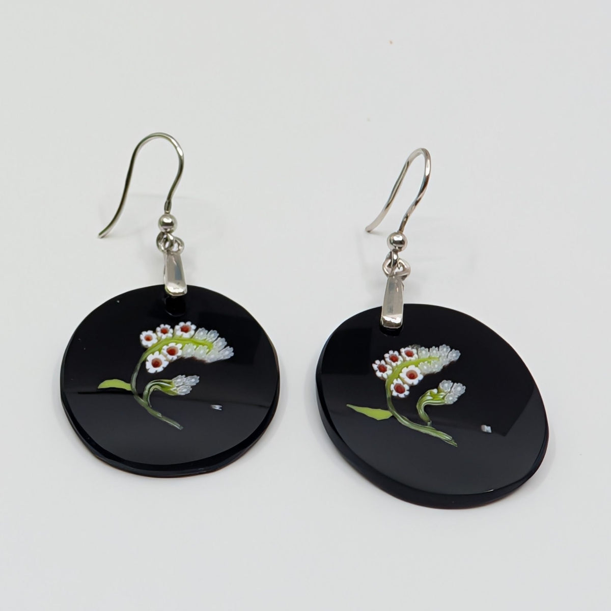 A pair of black round dangle earrings with silver fishhook wires, featuring a glass white, pink, and green flower design on the front, set against a plain light gray background.