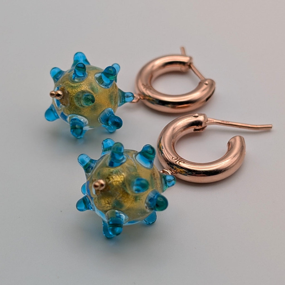 A pair of handmade drop earrings featuring textured, spiky translucent murano glass beads in blue and 24k gold, attached to rose gold hoop earrings, shown on a neutral light gray background.