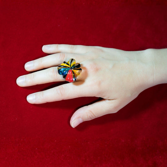 A hand wearing a colourful murano glass ring shaped like a stylised flower, set against a solid red background.