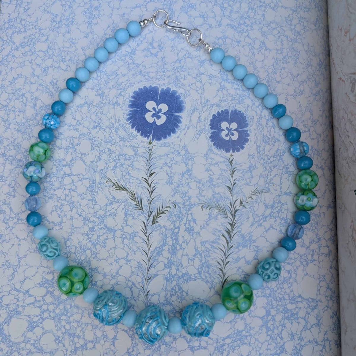 A beaded necklace with light blue, medium blue, and green beads, featuring both smooth round beads and textured carved accents, displayed on a blue and white floral patterned surface.