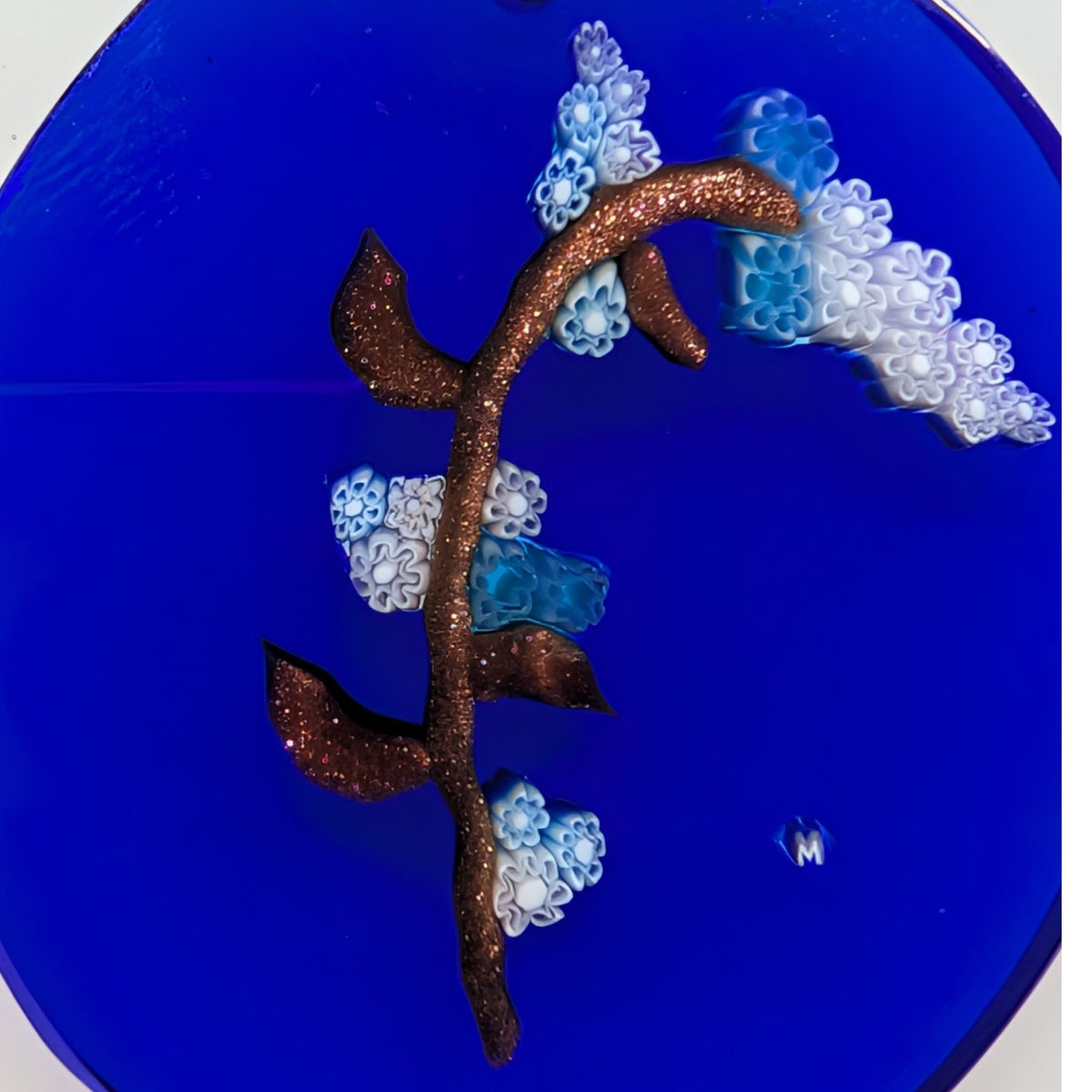 A close-up of a circular blue object featuring a raised gold branch with glittery texture and clusters of white and light blue floral or bubble-like details.