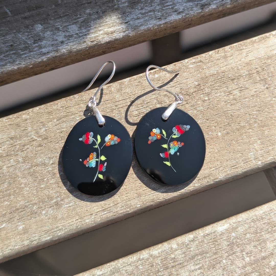 A pair of black oval dangle earrings with colorful butterfly and floral designs, hanging from silver fishhook wires on a weathered wooden surface.