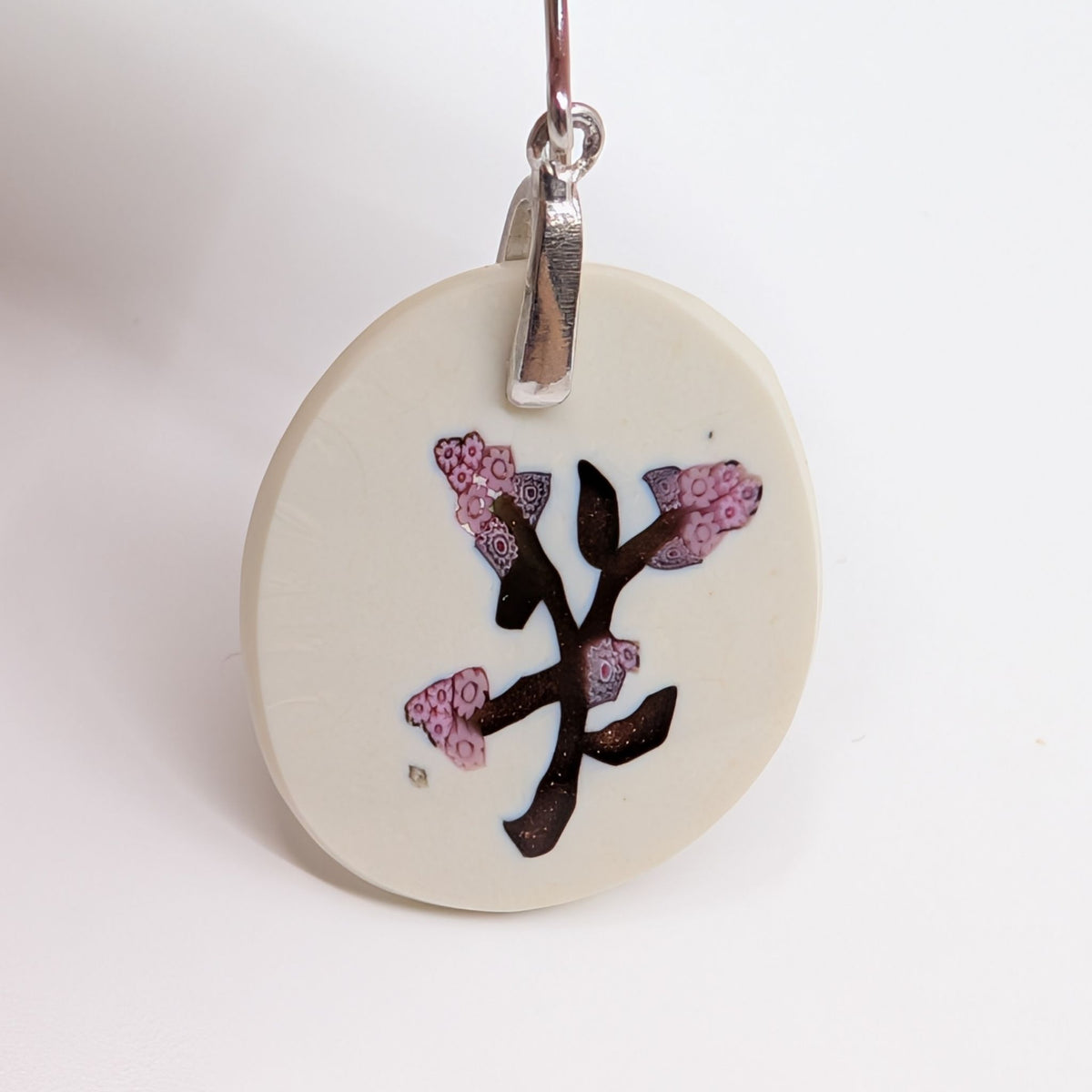 An oval millefiori earring with a cream-colored background featuring a gold branch design decorated with pink and purple textured floral details, suspended from a silver bail.