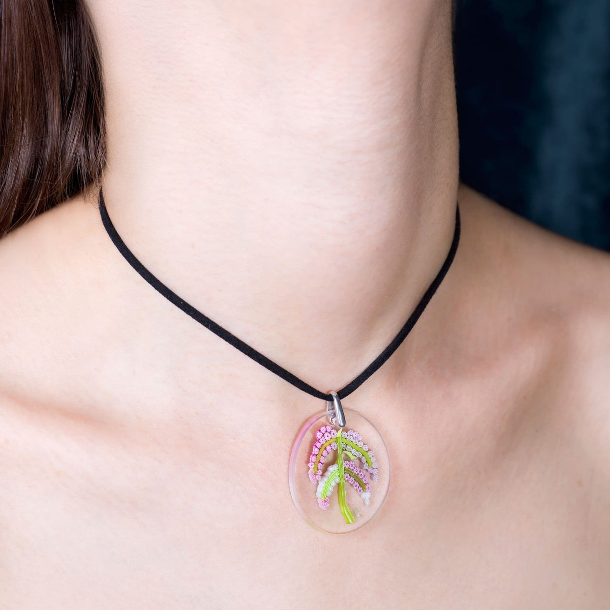 A close-up of a person's neck and collarbone wearing a black cord necklace with a clear oval pendant containing a small pink and green floral design.