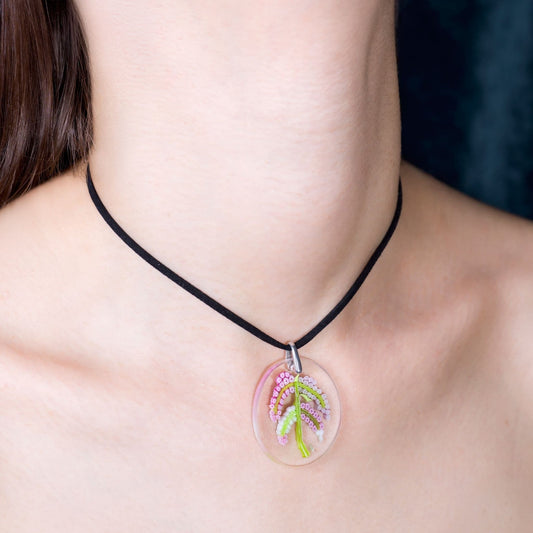 A close-up of a person's neck and collarbone wearing a black cord necklace with a clear oval pendant containing a small pink and green floral design.