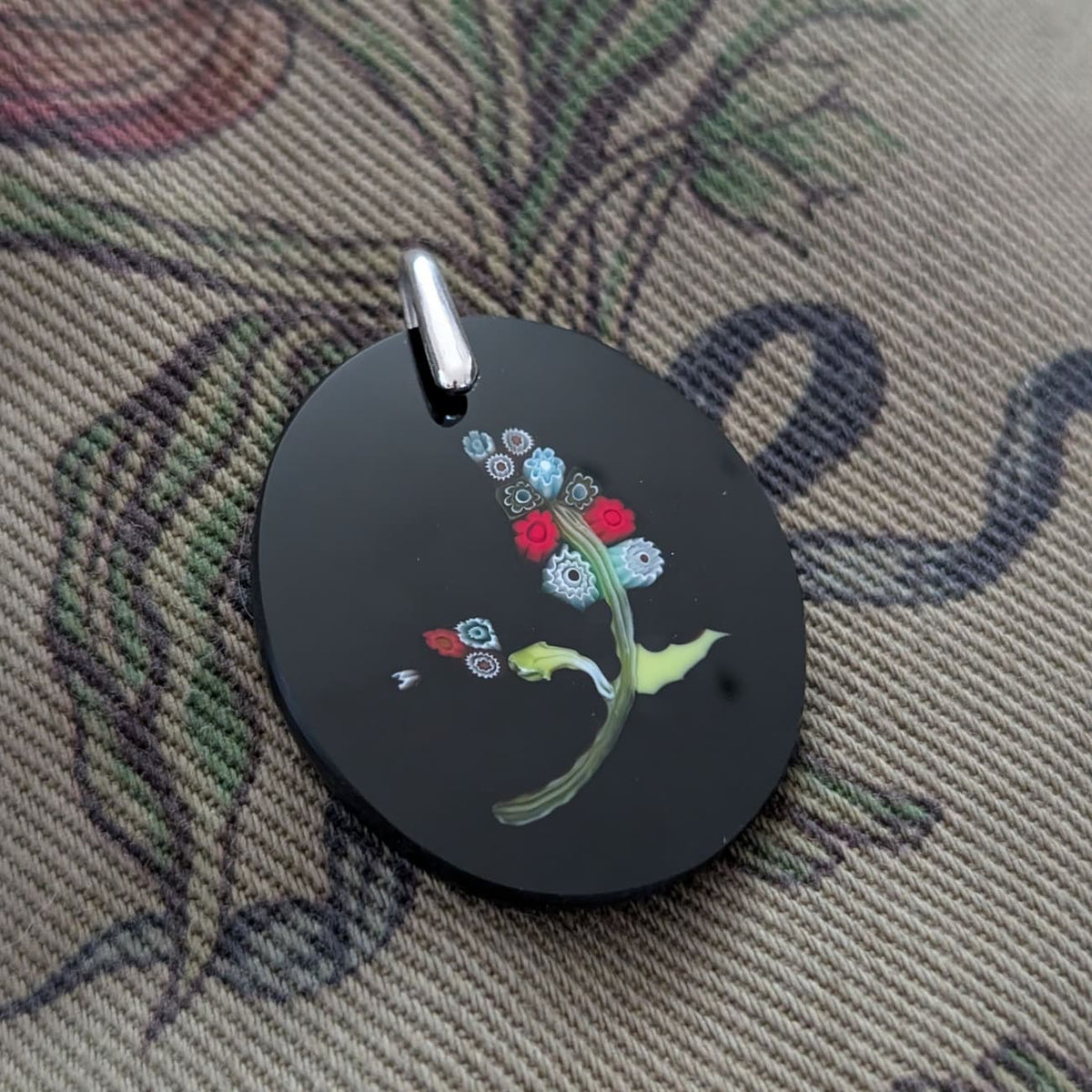 A round black pendant featuring a hand-painted floral design with red, blue, green, and yellow details, complete with a silver-tone metal bail, resting on a patterned fabric background.