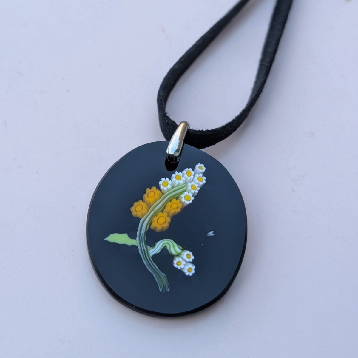 A round black pendant necklace featuring an illustration of yellow and white flowers with green stems, suspended from a black cord with a silver-toned metal bail.