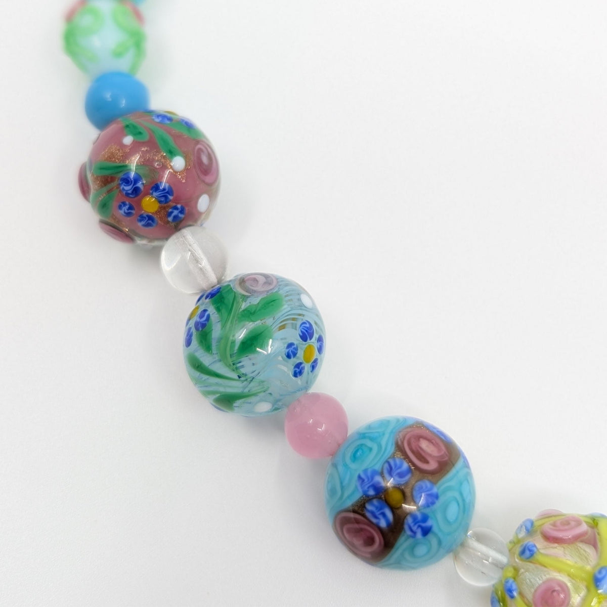 A close-up of a beaded strand featuring hand-painted glass lampwork beads in various colors and patterns, including blue, pink, green, and yellow, with clear glass spacer beads, on a plain white background.