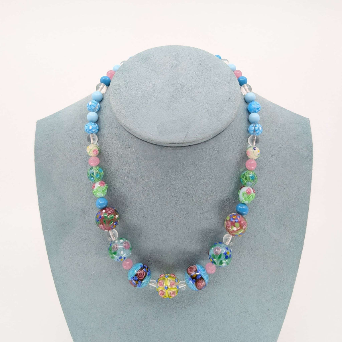 A colorful beaded necklace displayed on a light blue-gray jewelry bust against a plain white background. The necklace features a mix of blue, pink, green, and multicolored patterned glass beads, including clear faceted beads.