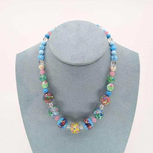 A colorful beaded necklace displayed on a light blue-gray jewelry bust against a plain white background. The necklace features a mix of blue, pink, green, and multicolored patterned glass beads, including clear faceted beads.