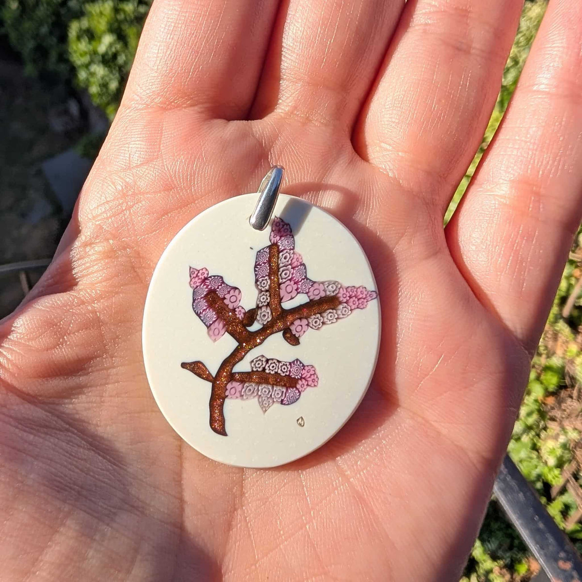 A hand holding a round white pendant featuring a cherry blossom branch design with pink and purple blossoms, photographed outdoors with blurred greenery in the background.