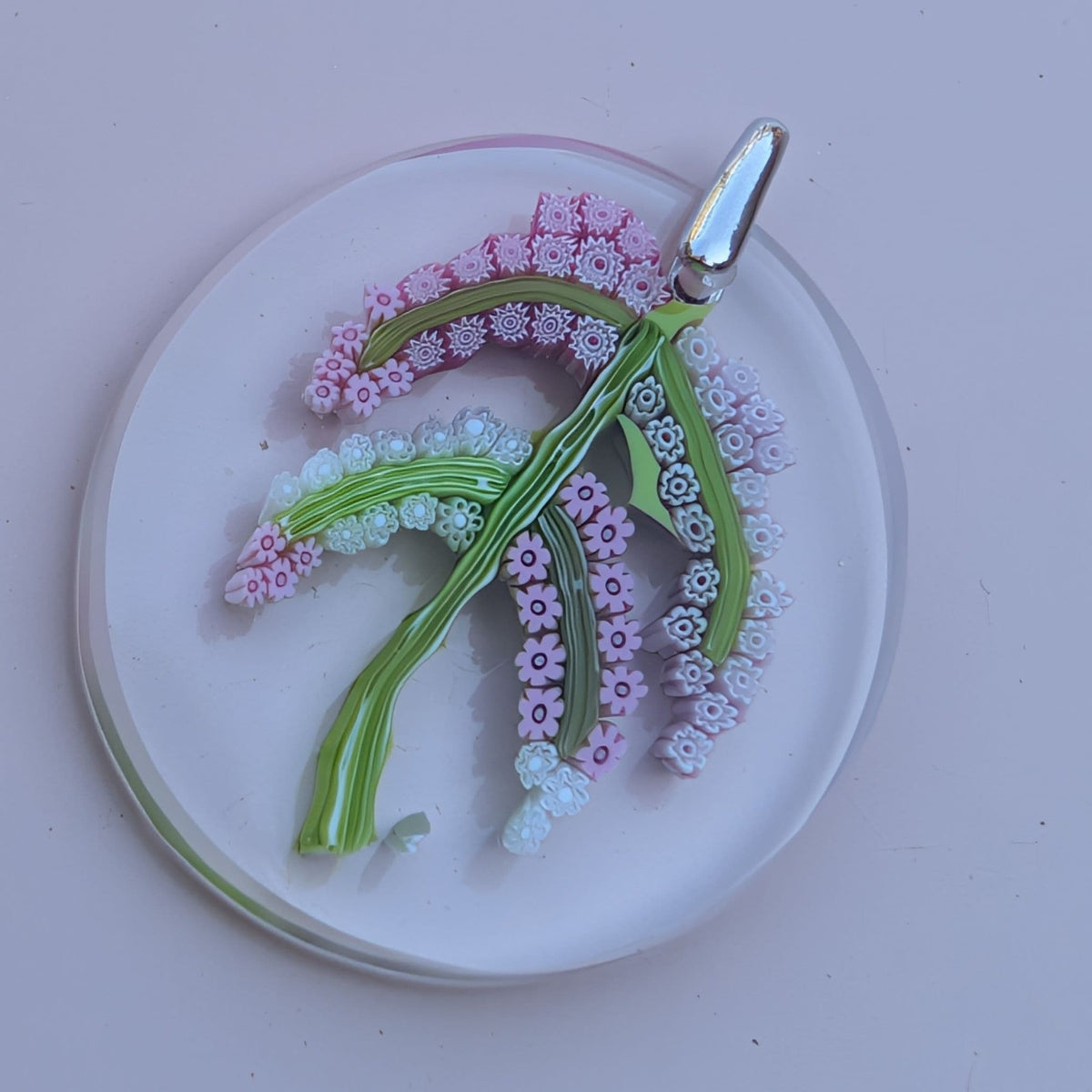 A circular glass pendant featuring a detailed floral vine design in shades of pink, green, and white, with a polished silver bail at the top, displayed on a light pink background.