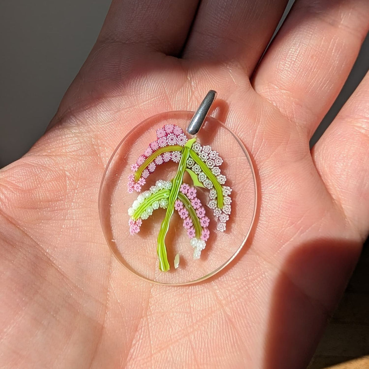 A close-up of a hand holding a round clear murano glass pendant featuring a delicate floral design in pink, white, and green, with a silver metal loop at the top.