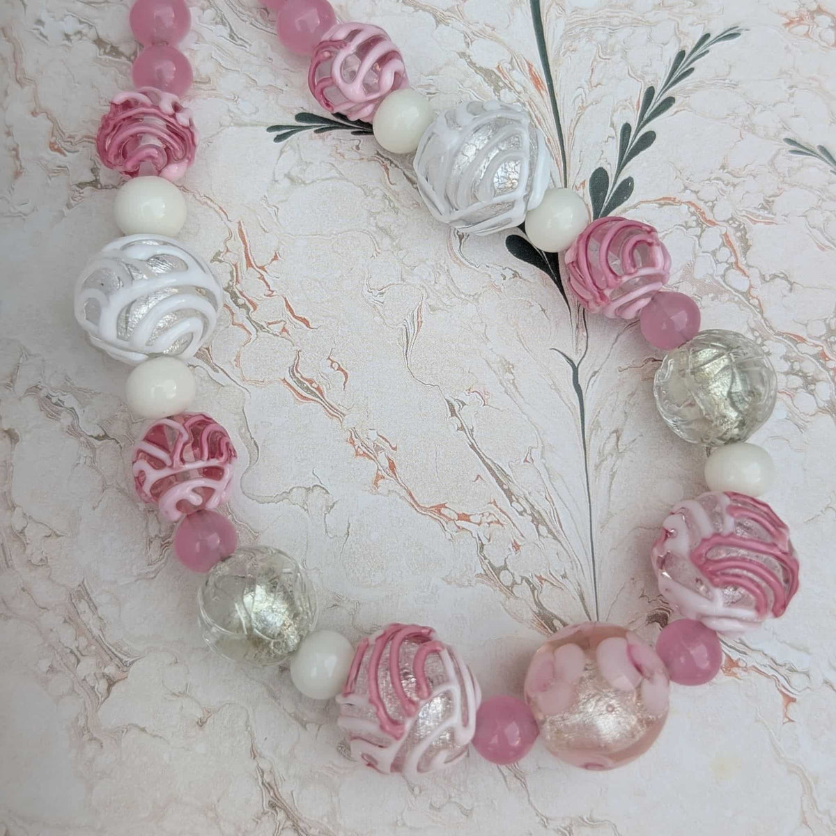 A handmade beaded necklace featuring pink and white patterned glass beads, white pearl-like beads, and textured clear glass beads, displayed on a light marble-patterned background.