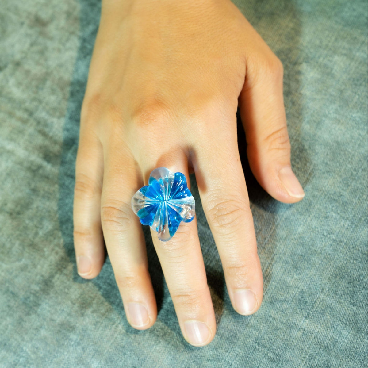 A hand wearing a large, sculptural flower-shaped ring made of transparent blue and clear glass.