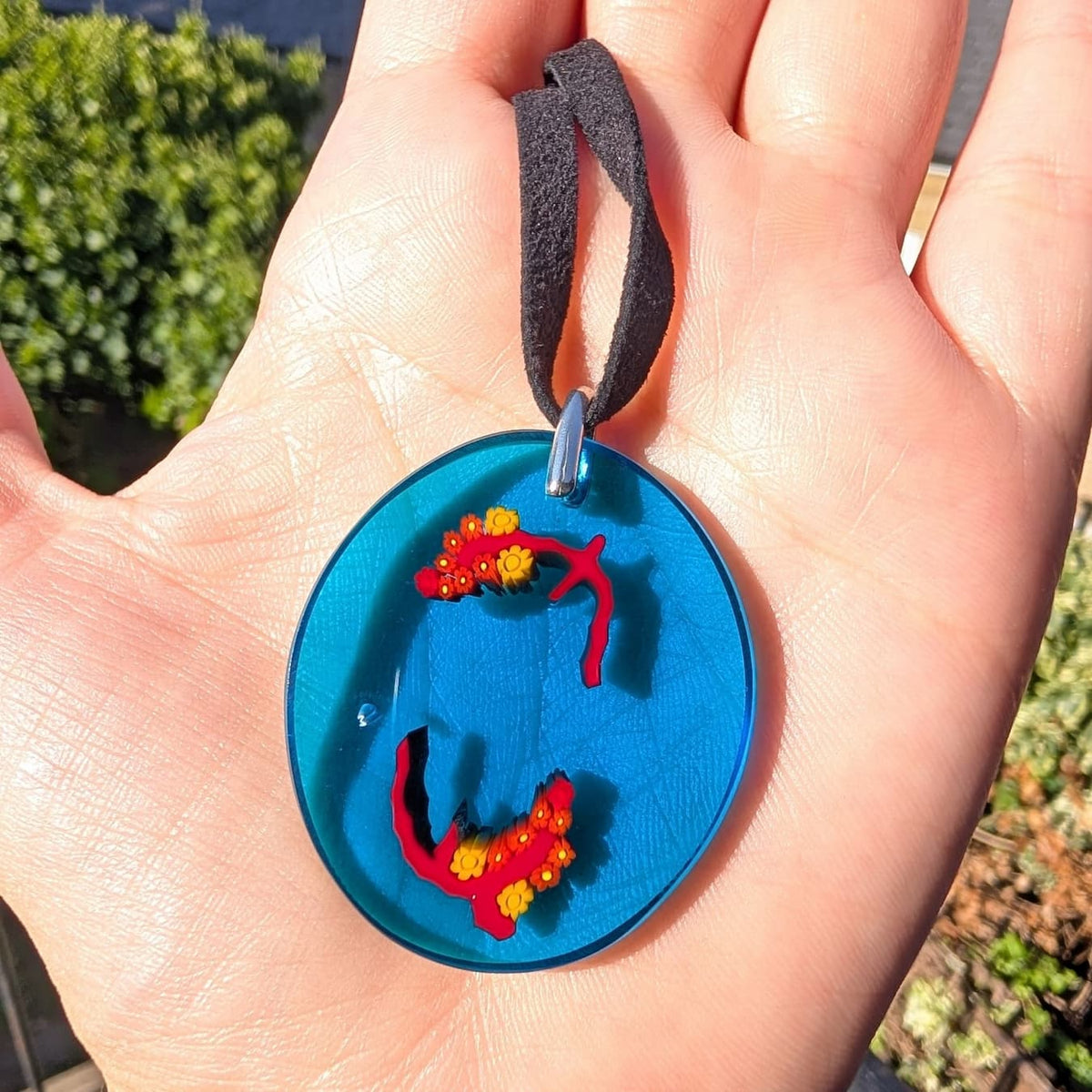 A close-up of a hand holding a round blue pendant necklace with red and yellow floral details and a black cord, photographed outdoors with blurred greenery in the background.