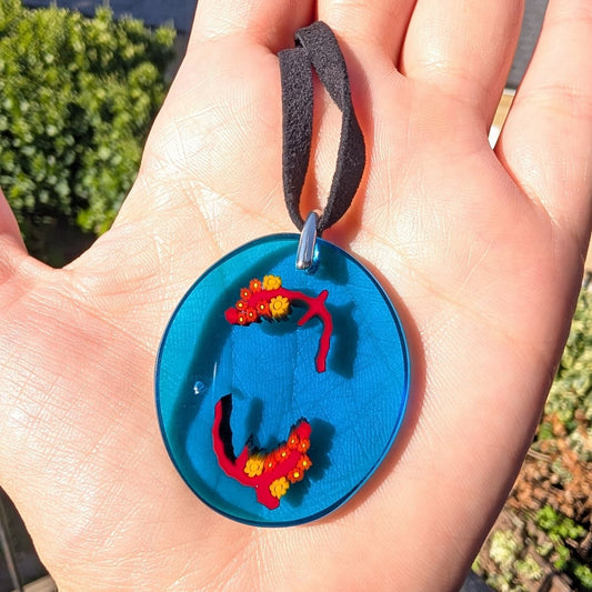 A close-up of a hand holding a round blue pendant necklace with red and yellow floral details and a black cord, photographed outdoors with blurred greenery in the background.