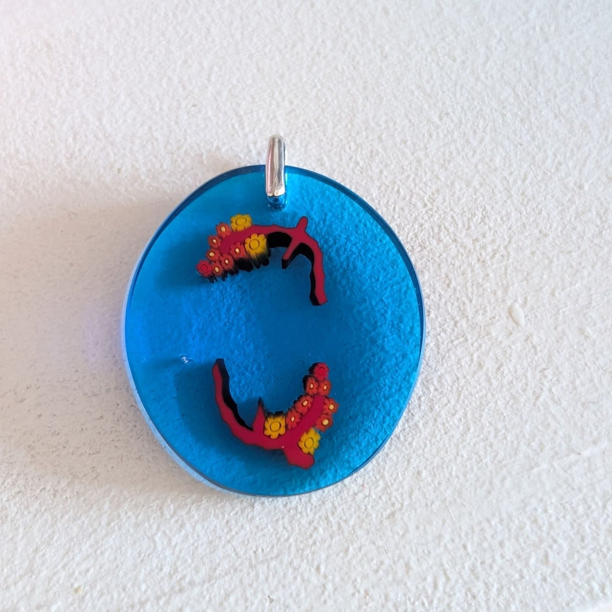 A round, translucent blue pendant featuring two small, red and yellow floral bird charms, displayed on a textured white background.