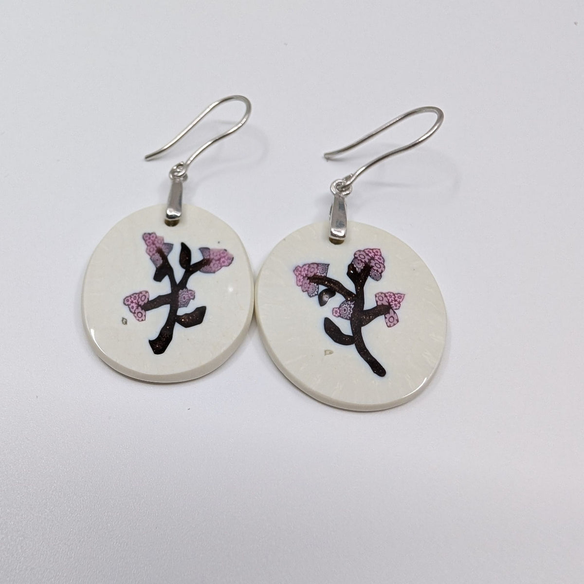 A pair of handmade dangle earrings featuring round, off-white glass earrings decorated with a black branch motif and pink cherry blossoms, suspended from silver fishhook wires on a plain white background.